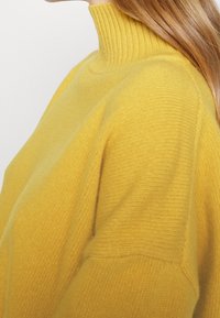 Kobieta w jasnożółtym, żebrowanym swetrze z golfem, z długimi, prostymi, jasnobrązowymi włosami widocznymi z boku.