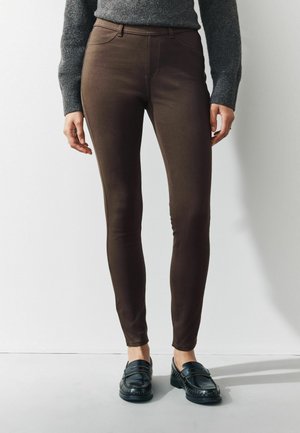 Braune enge Jeans aus Stretchstoff, mit hohem Bund und vorderen Taschen, kombiniert mit schwarzen Slippern. Glatte Textur, taillierte Passform.
