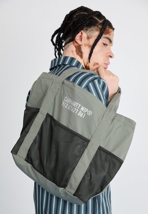 Szürke tote táska, fekete hálós aljjal és oldalpanelekkel. Carhartt WIP logóval rendelkezik. Vállon viselve, csíkos inggel.