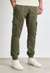 Olive green cargo pants with elastic cuffs and pockets on the thighs, made of durable fabric, paired with white sneakers.