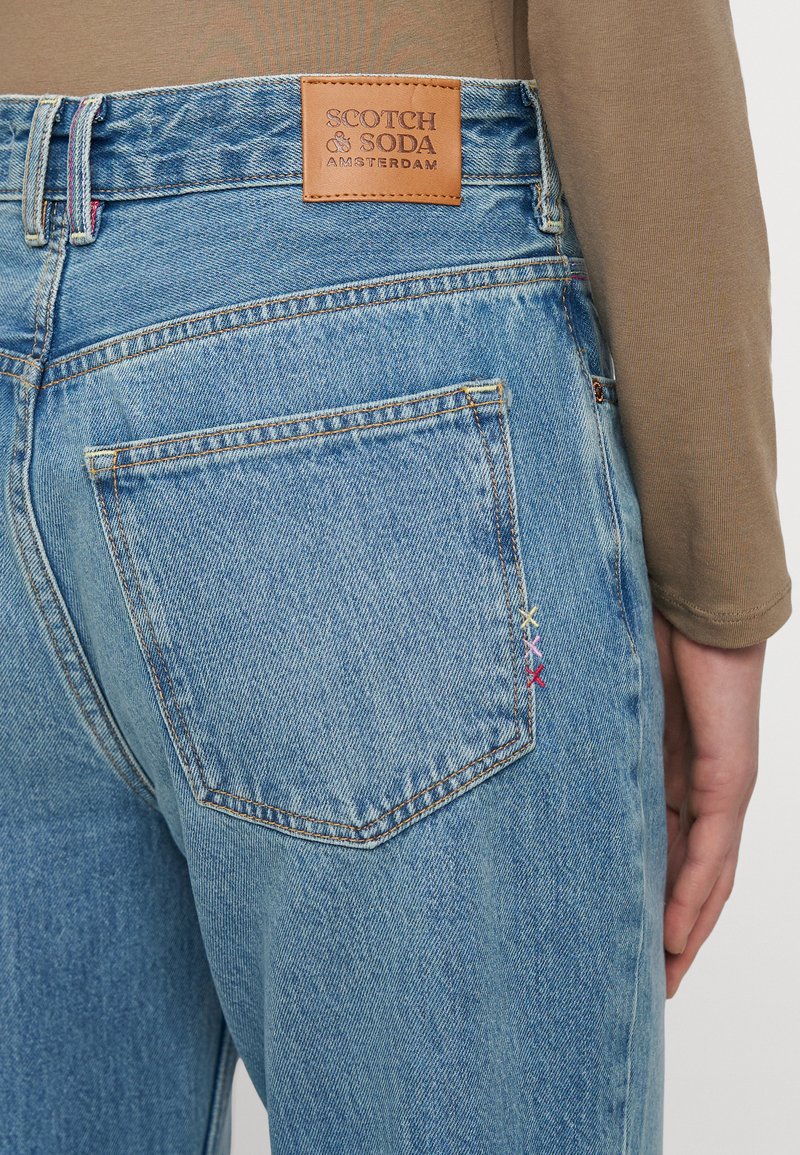 Jeans de mezclilla azul claro con una etiqueta de marca de cuero, dos bolsillos traseros y acentos bordados coloridos. Diseño clásico de cinco bolsillos.