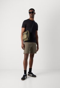 Sort t-shirt, olivengrønne shorts og sorte sneakers med hvide striber; bærer en lille olivengrøn crossover-taske; iført solbriller.