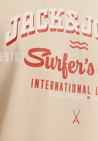 Beige katoenen t-shirt met gedurfde rode en witte tekst: "JACK&JONES Surfer's," met golven en symbolen eronder gedrukt. Gladde textuur.