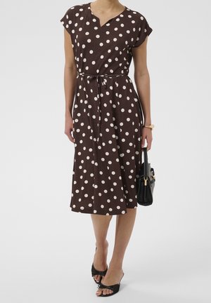 Mujer que lleva un vestido midi marrón con lunares blancos, tacones abiertos negros y lleva un bolso negro en la mano derecha.