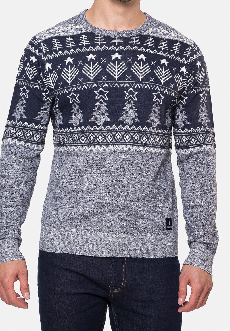 Homme portant un pull gris et bleu marine avec des motifs de sapin de Noël, d'étoiles et géométriques, assorti à un jean bleu foncé.