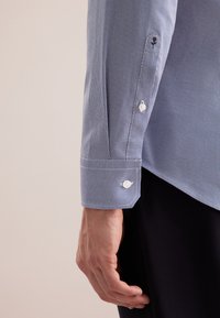 Rechterarm in een lange blauwe shirt met patroon en witte knopen, hand ontspannen langs de zijkant, en zwarte broek zichtbaar.