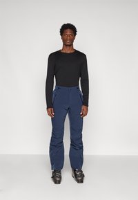 Zwarte lange mouwen top gecombineerd met marineblauwe skibroeken, met ritssluitingen en een gladde, duurzame stof. Zwarte skiboots maken de outfit af.