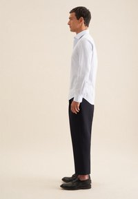 Camicia bianca a maniche lunghe con colletto button-down, abbinata a pantaloni scuri e scarpe nere. Il tessuto appare liscio con una vestibilità su misura.