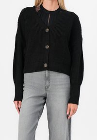 Cardigan negro de punto con escote en V, mangas largas y cierre de cinco botones. Lleva puesto sobre una blusa negra y jeans gris claro. Tejido texturizado.
