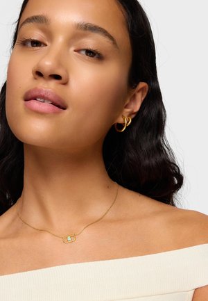 Młoda kobieta o średnim odcieniu skóry, nosząca małe złote kolczyki w kształcie kółek oraz delikatny złoty naszyjnik z owalu połączonym ogniwem.