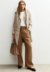 Manteau en laine beige clair avec des revers surdimensionnés, top blanc ajusté, pantalons évasés marron avec cordon de serrage, sac tote assorti et ballerines noires.
