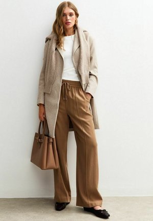 Manteau en laine beige clair avec des revers surdimensionnés, top blanc ajusté, pantalons évasés marron avec cordon de serrage, sac tote assorti et ballerines noires.