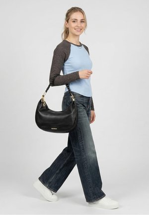 Mladá žena kráčející, oblečená v světle modrém a šedém tričku s dlouhým rukávem, širokých tmavých džínách, bílých teniskách, nesoucí na předloktí černou koženou kabelku.