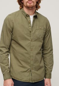 Camisa de manga larga verde oliva hecha de algodón, con cuello abotonado, bolsillo en el pecho y botones de latón a lo largo de la tapeta delantera.
