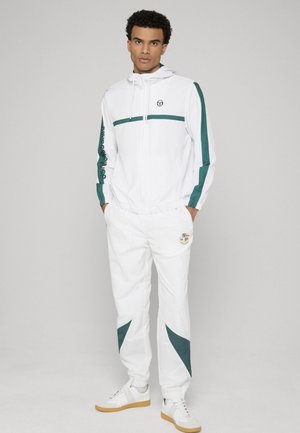 Uomo in piedi con le mani in tasca, indossa una tuta bianca con strisce verdi e sneakers bianche, sullo sfondo grigio chiaro.