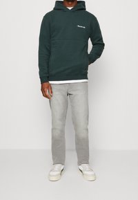 Un hoodie vert foncé avec une poche kangourou, présentant un texte brodé blanc sur la poitrine. Associé à un jean gris clair et des baskets blanches.