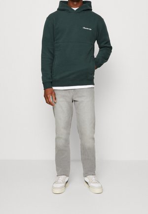 Felpa verde scuro con tasca a marsupio, caratterizzata da testo ricamato bianco sul petto. Abbinata a jeans grigi chiari e sneakers bianche.