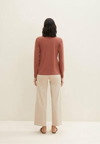 Frau mit schulterlangem dunklem Haar steht mit dem Rücken zur Kamera, trägt ein langärmliges, rostfarbenes Oberteil, beige Hose mit weitem Bein und braune Sandalen.