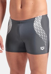 Costumi da bagno grigi scuri realizzati in materiale liscio, con un motivo geometrico bianco sui lati e un logo sull'orlo.