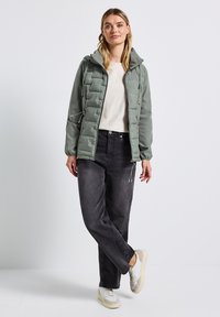 Gewatteerde lichtgroene jas met capuchon, gecombineerd met een beige trui en donkergrijze jeans. Witte en grijze sneakers maken de outfit compleet.
