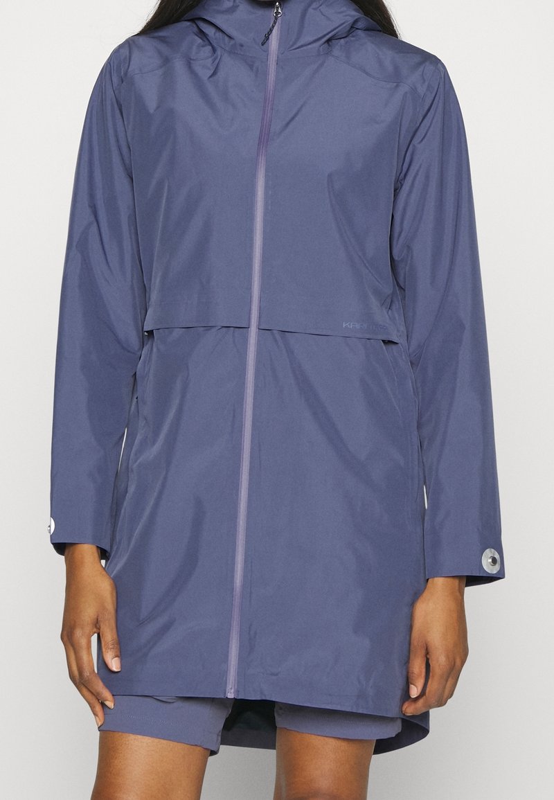 Femme portant un imperméable violet à manches longues, avec capuche, fermeture éclair à l'avant et poignets boutonnés.