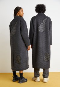 Două persoane îmbrăcate în paltoane lungi, gri închis, din lână, cu accente de țesătură texturată pe spate, care prezintă forme și modele unice.