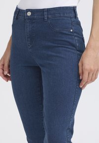 Jeans en denim bleu foncé avec une coupe slim, poches avant, fermeture à bouton en métal argenté et détails de couture discrets le long des coutures.