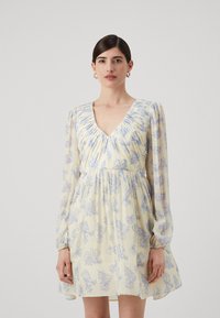 Kukullinen mekko vaaleankeltaisena sinisillä kuvioilla, jossa on V-pääntie, rypytetty yläosa ja läpikuultavat pitkät hihat.