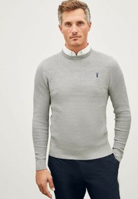 Pull en tricot gris avec un col rond, des poignets côtel�és et un petit logo bleu sur le côté gauche de la poitrine, superposé sur une chemise blanche à col.