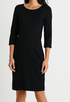 Femme portant une robe noire longueur genou avec des manches trois-quarts et un décolleté arrondi, se tenant devant un fond uni.