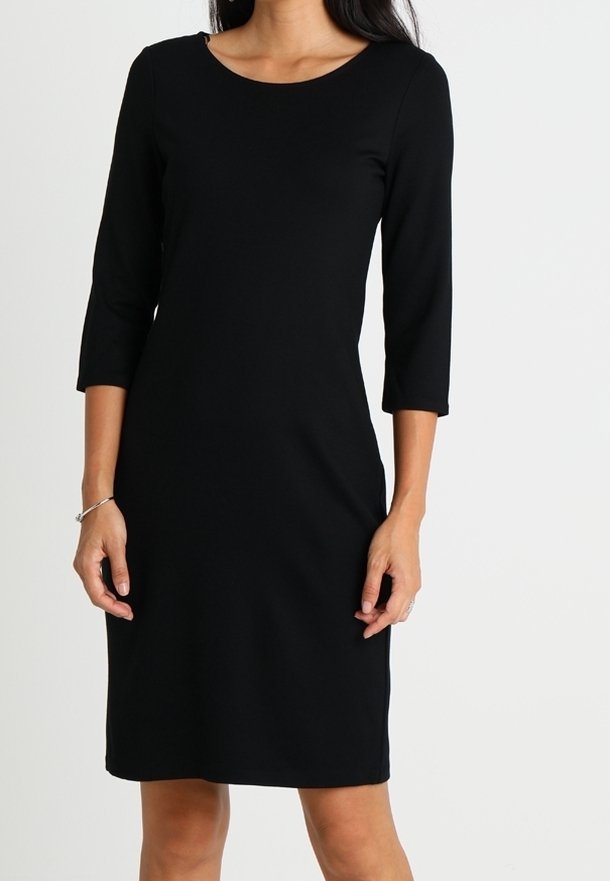 Femme portant une robe noire longueur genou avec des manches trois-quarts et un décolleté arrondi, se tenant devant un fond uni.