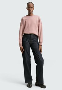 Sudadera rosa claro con hombros caídos, combinada con jeans de mezclilla oscuros de pierna ancha que tienen una cintura alta y corte recto. Zapatos negros gruesos.