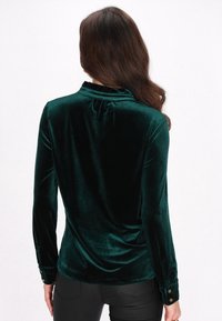 Mørkegrøn fløjl blouse med høj krave, lange ærmer med knoplukninger og en diskret samling detalje bagpå.