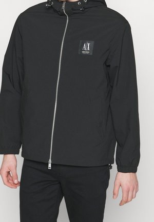 Chaqueta ligera negra con capucha, cierre frontal de cremallera, puños elásticos y parche con logo en la parte superior izquierda del pecho. Textura de tela suave.
