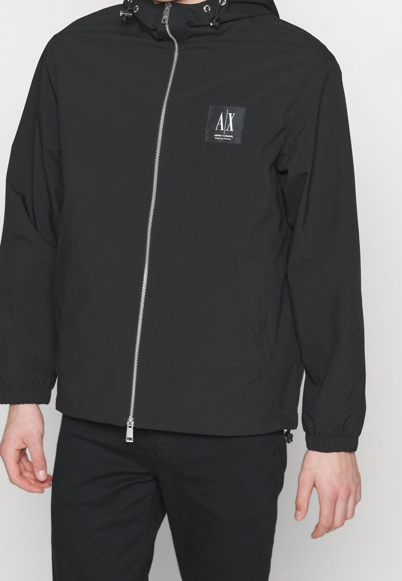 Svart lättviktjacka med huva, framatgångdragkedja, elastiska ärmslut och logotyptryck på övre vänstra bröstet. Slät tygstruktur.