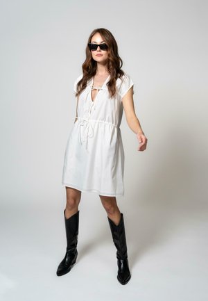 Robe en coton blanche avec des manches courtes et un détail noué au col, associée à des bottes noires hautes jusqu'aux genoux. Coupe simple et décontractée.