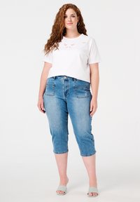 Witte cropped t-shirt met decoratieve borduursels, gecombineerd met lichtblauwe denim capri-broeken met voor- en achterkant zakken, en lichtgrijze instapschoenen.