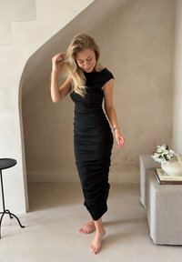 Rochie neagră, ajustată, până la genunchi, cu mâneci scurte și detalii pliate pe corp. Purtați descult într-un interior cu tonuri neutre.