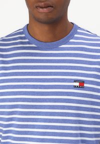 Riba sinine ja valge t-särk ümmarguse kaelusega ja lühikeste varrukatega; vasakul rinnapiiril on väike "Tommy Jeans" logo.