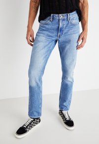 Jeans in denim blu chiaro con vestibilità dritta e texture sbiadita, caratterizzati da un tradizionale design a cinque tasche e chiusura frontale con bottone. Abbinati a sneaker con motivo bianco e nero.