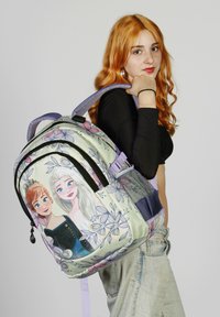 Mochila con una tela verde claro con patrones florales y un gráfico de dos personajes. Correas ajustables moradas y cremalleras negras.