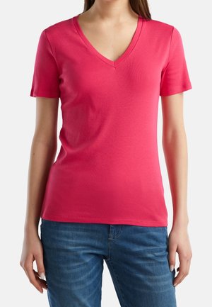 Kobieta w jasnoróżowej koszulce z krótkim rękawem i dekoltem w serek oraz niebieskich dżinsach, stojąca na tle jednolitego białego tła.