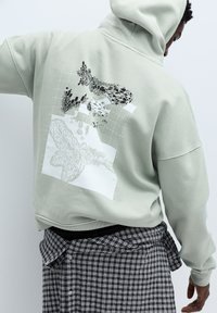 Lichtgroene hoodie met een grafisch ontwerp met zwart-witte abstracte patronen op de achterkant. Zachte katoenen stof met een ontspannen pasvorm.