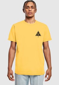 Gelbes Baumwoll-T-Shirt mit kurzen Ärmeln, versehen mit einer schwarzen dreieckigen Grafik mit einem Auge auf der linken Brust. Klassischer Rundhalsausschnitt.