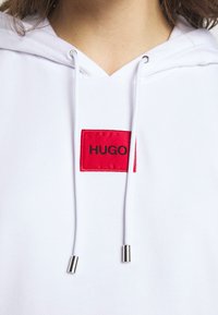 Sudadera blanca con un parche rectangular rojo con la palabra "HUGO" en negro. Puntas de cordón metálicas en tono plateado. Tejido suave y texturizado.