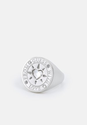 Zilveren ring met een hartvormige kristal in het midden, zonnestralen ontwerp, vier kleine kristallen en de woorden "FROM GUESS WITH LOVE" rond de rand.