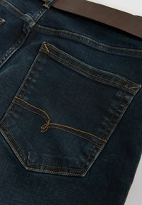 Jeans de mezclilla azul oscuro con un solo bolsillo trasero con costuras amarillas y una cintura de cuero marrón. La tela tiene una textura suave.