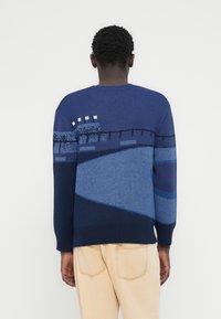 Άτομο που φοράει πουλόβερ με σχέδιο φράχτη σε σκούρο μπλε και μπλε χρώμα, σε συνδυασμό με ανοιχτό μπεζ παντελόνι, με την πλάτη γυρισμένη προς τα πίσω, μπροστά σε απλό φόντο.