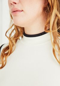 Close-up of a person’s lower face and neck with light skin, wavy red hair, wearing a white shirt with a black collar.