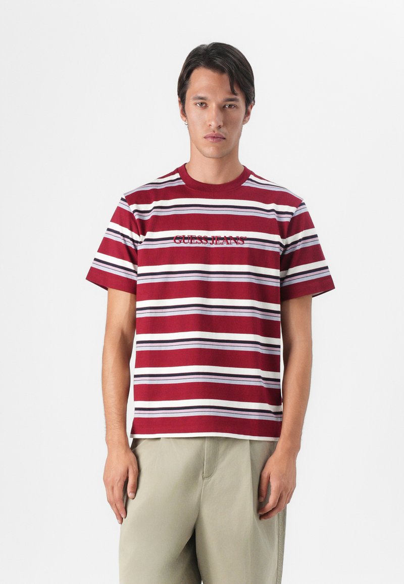 Punane ja valge triibuline T-särk lühikese varrukaga, millel on burgundis värvitud "GUESS JEANS". Ümmargune kaelus, lõdvestunud sobivus.